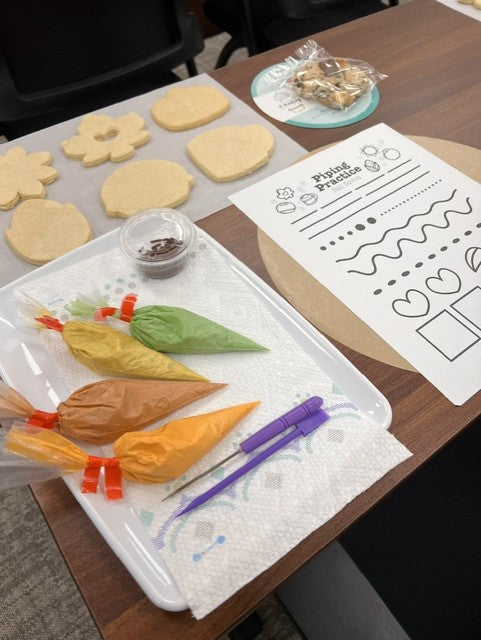 A baking or craft activity setup on a wooden table, featuring a white tray with piping bags filled with orange, yellow, green, and brown icing, a purple tool, a printed worksheet labeled 'Piping Practice,' and several pale dough cutouts on a sheet.