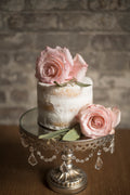 Small white cake with pink roses on a decorative stand against a brick wall.