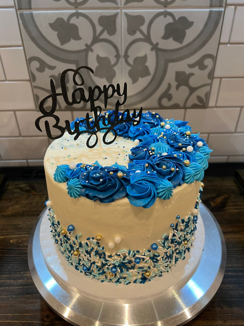 Two-tier birthday cake with blue flowers and 'Happy Birthday' topper on a tiled wall background
