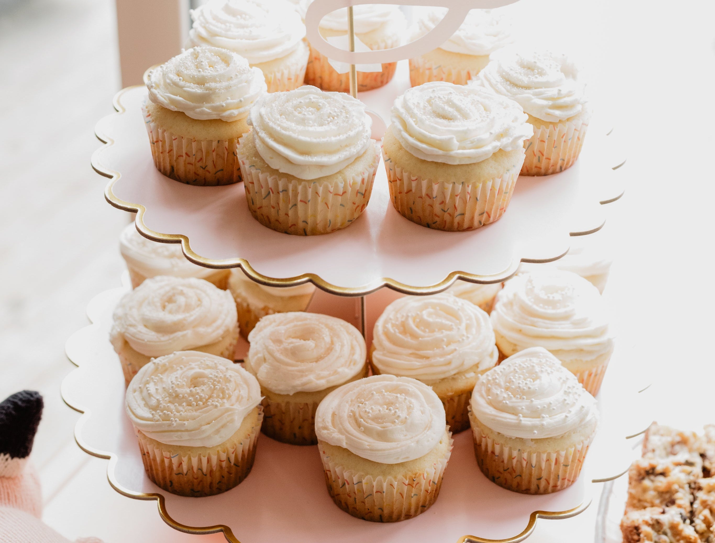 A two-tiered pink cupcake stand with gold scalloped edges, filled with frosted cupcakes